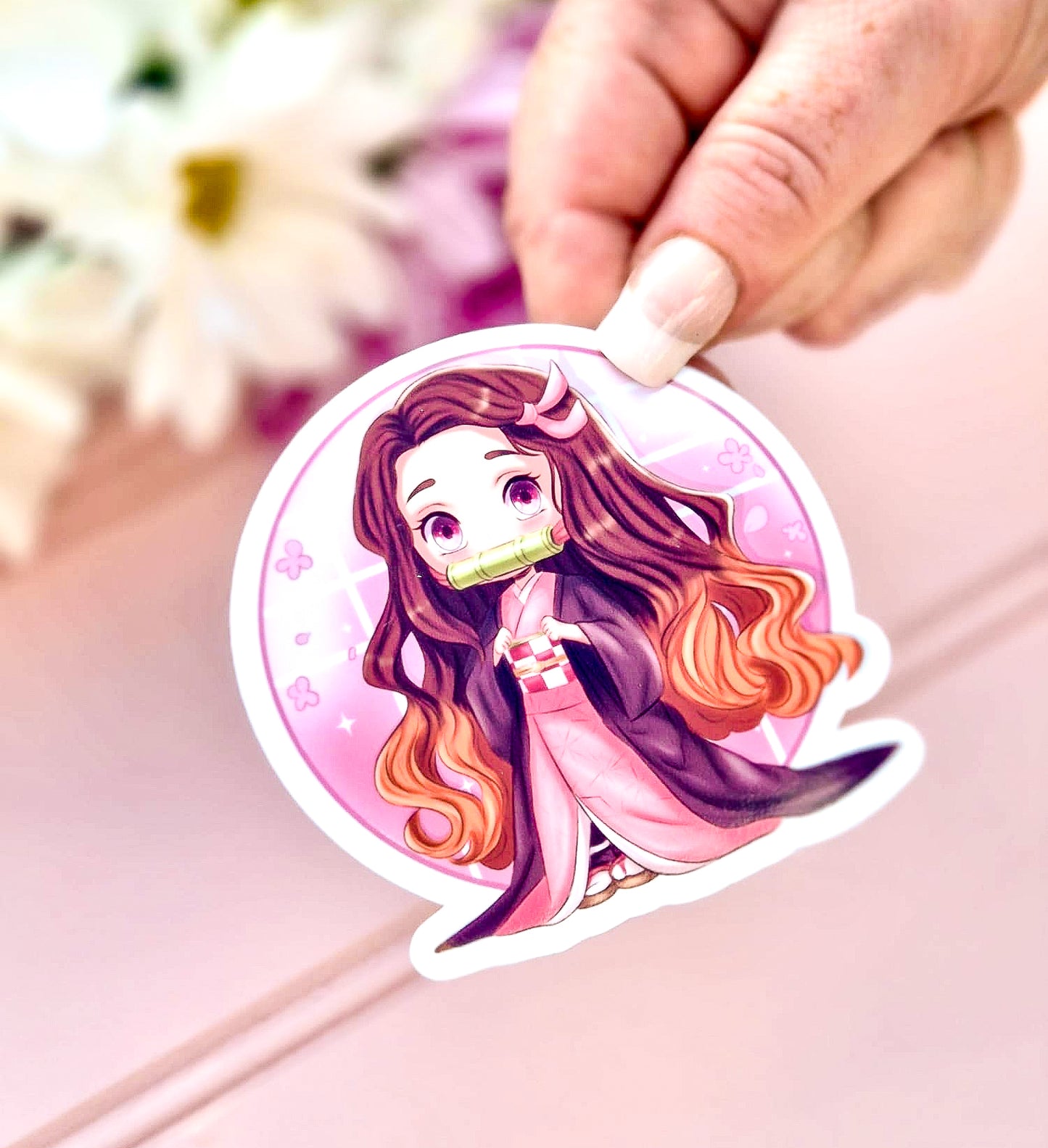 Sticker of nezuko demon slayer character with long hair held by a hand on a pink surface.