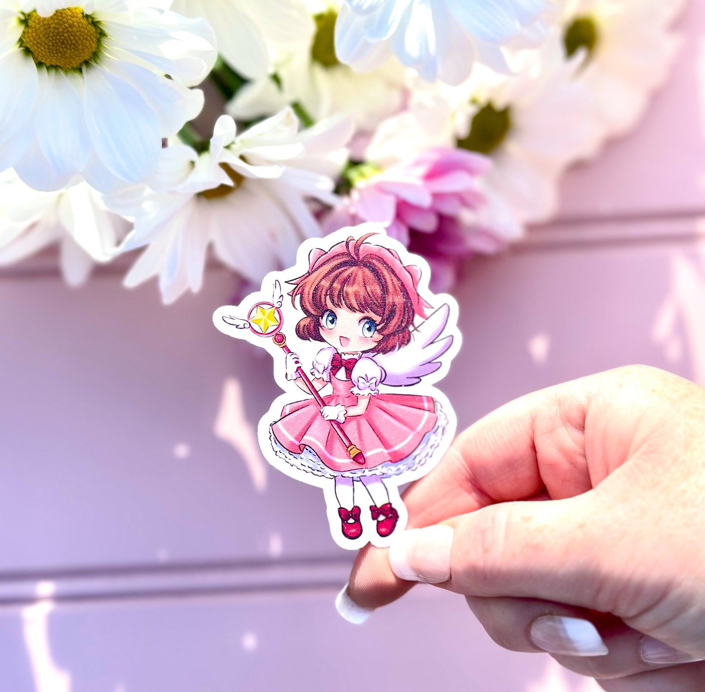 Hand holding a sticker of an anime character sakura from cardcaptors with flowers in the background
