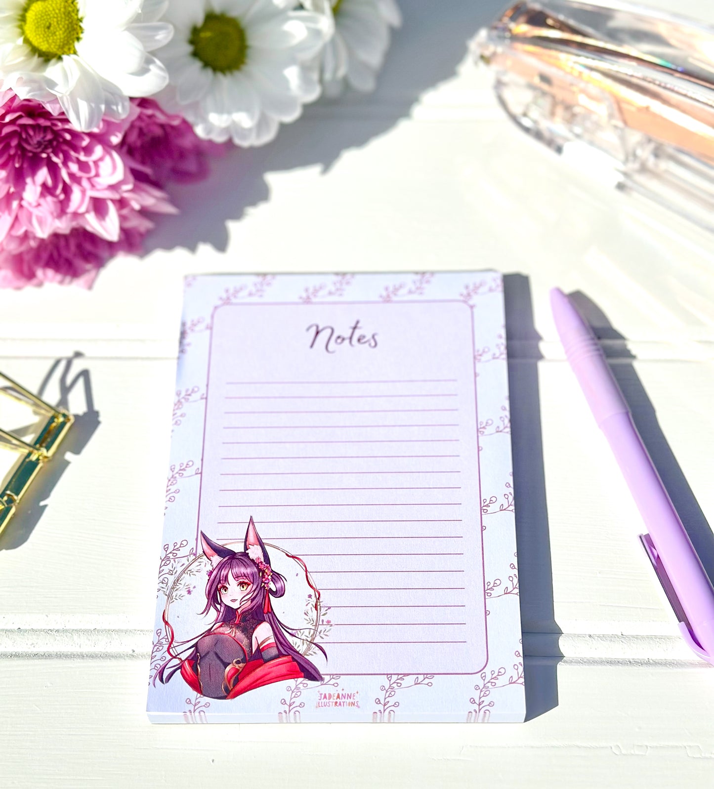 Notepad with anime kitsune character design on a table with flowers and a pen.