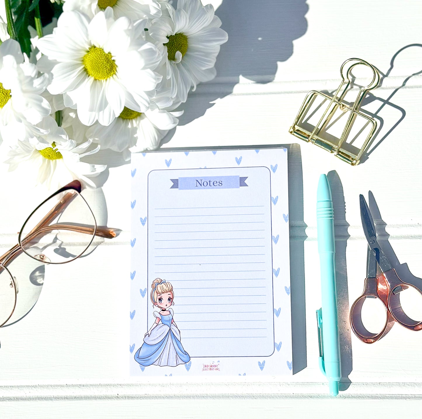 Notepad with chibi princess design, glasses, flowers, pen, and scissors on a white surface