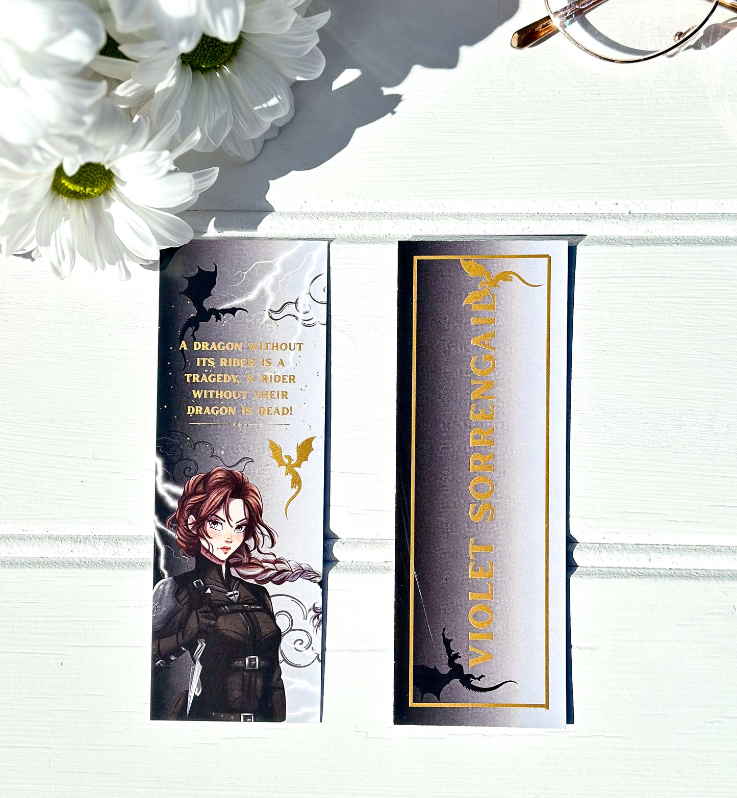 Bookmarks with Violet sorrengail from fourth wing and text on a white surface with flowers and glasses.