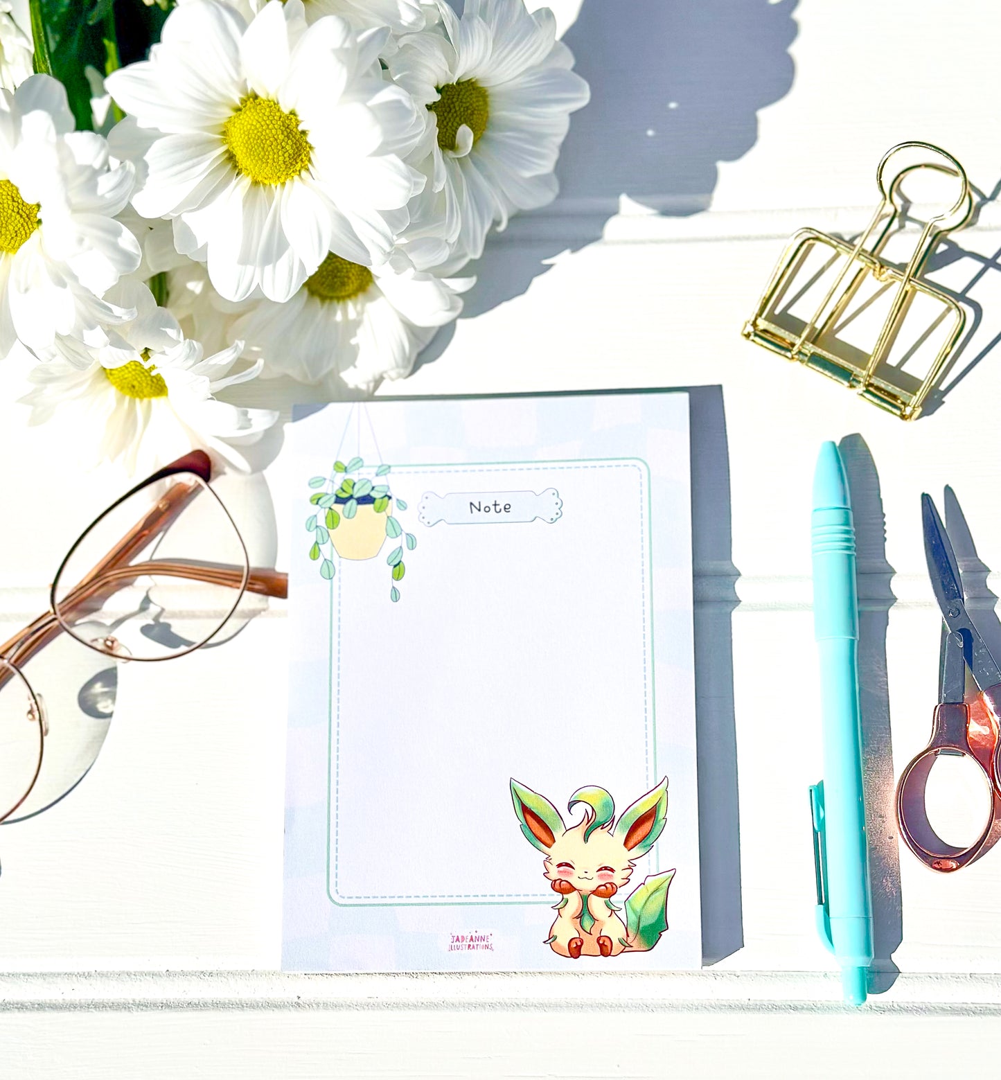 Notepad with kawaii leafeon illustration, glasses, flowers, pen, and scissors on a white surface