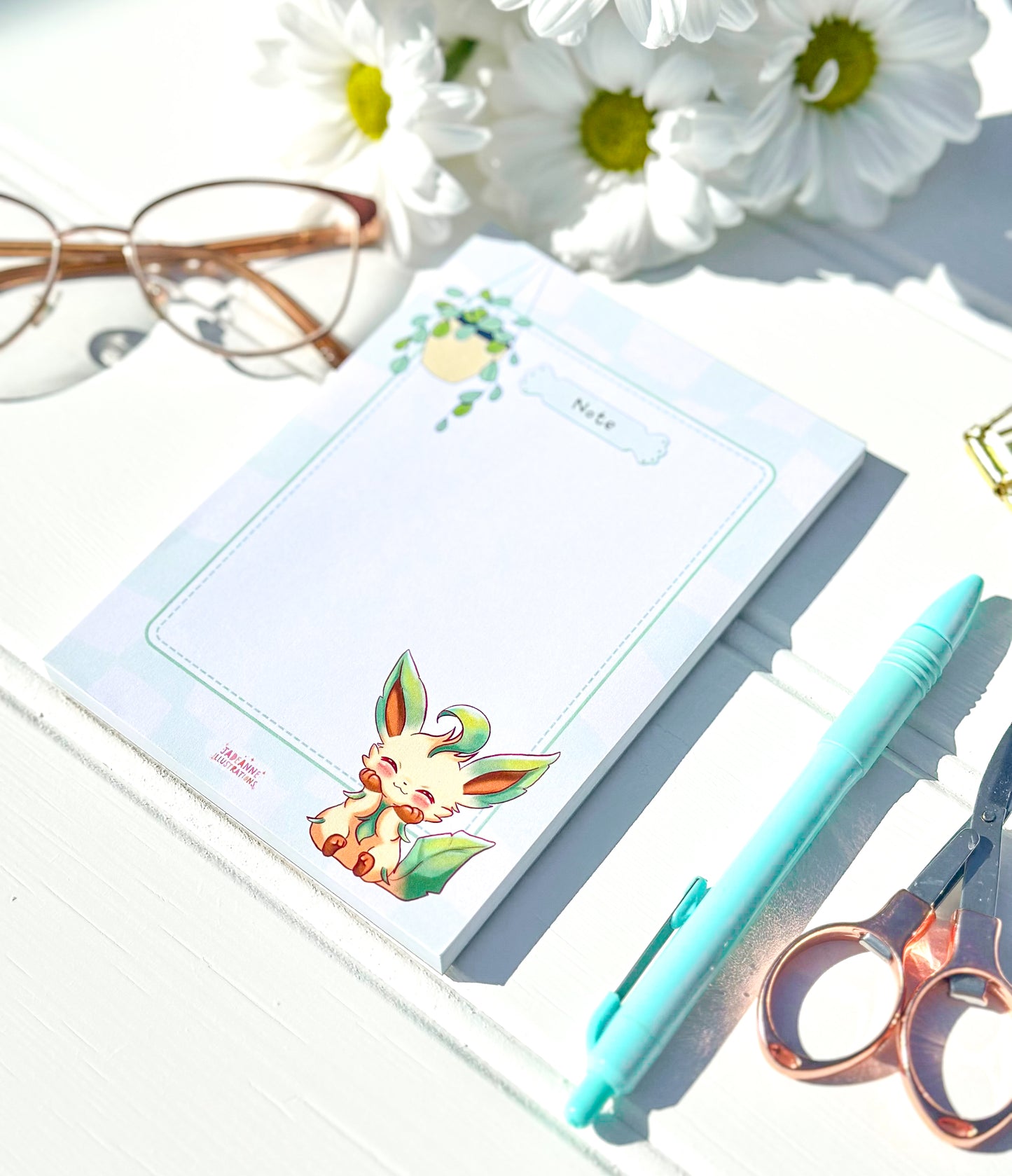 Notepad with kawaii pokemon character design, pen, scissors, and glasses on a light surface with flowers.