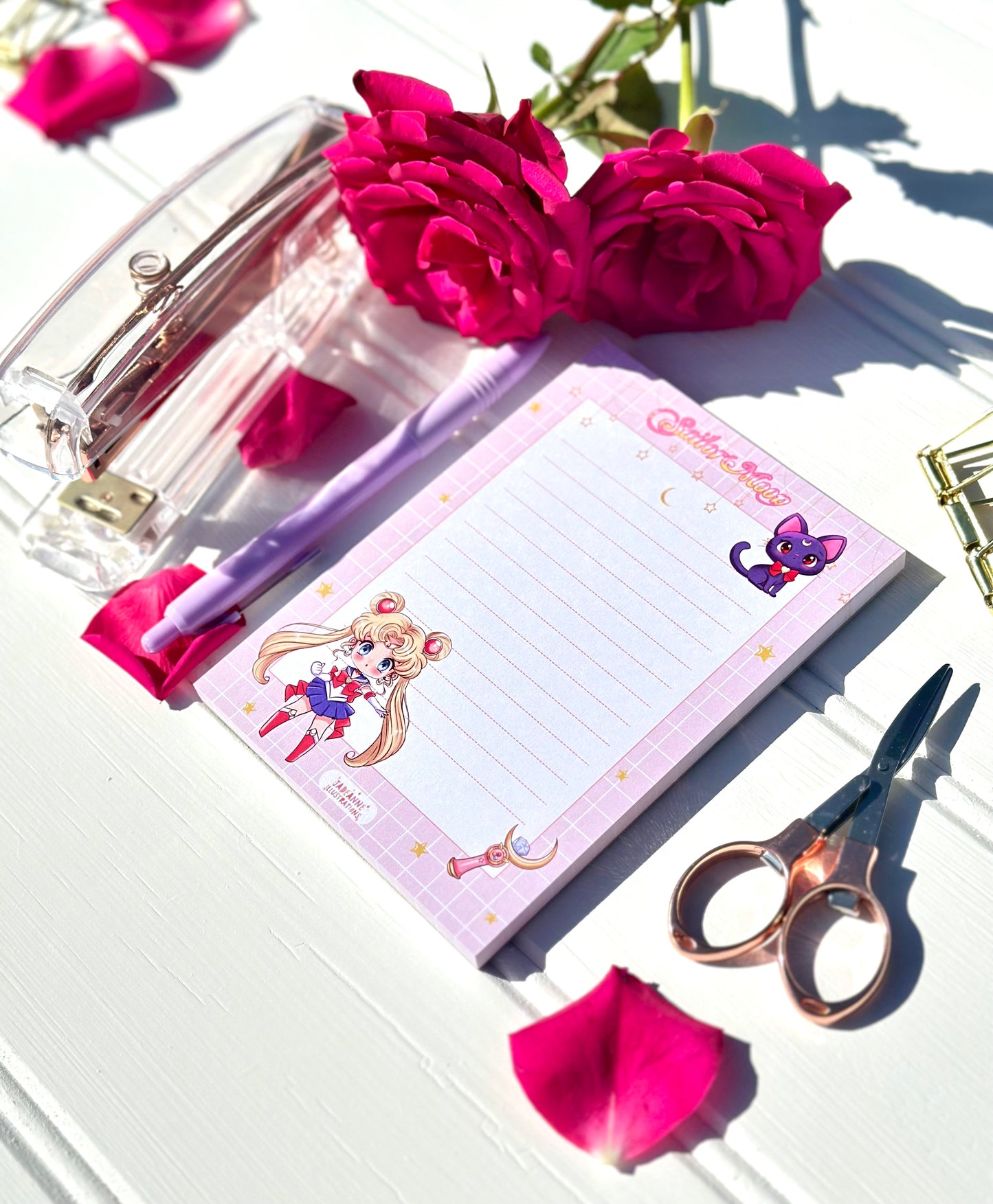 Notebook with Sailor Moon design, pen, and scissors on a white surface with pink flowers.