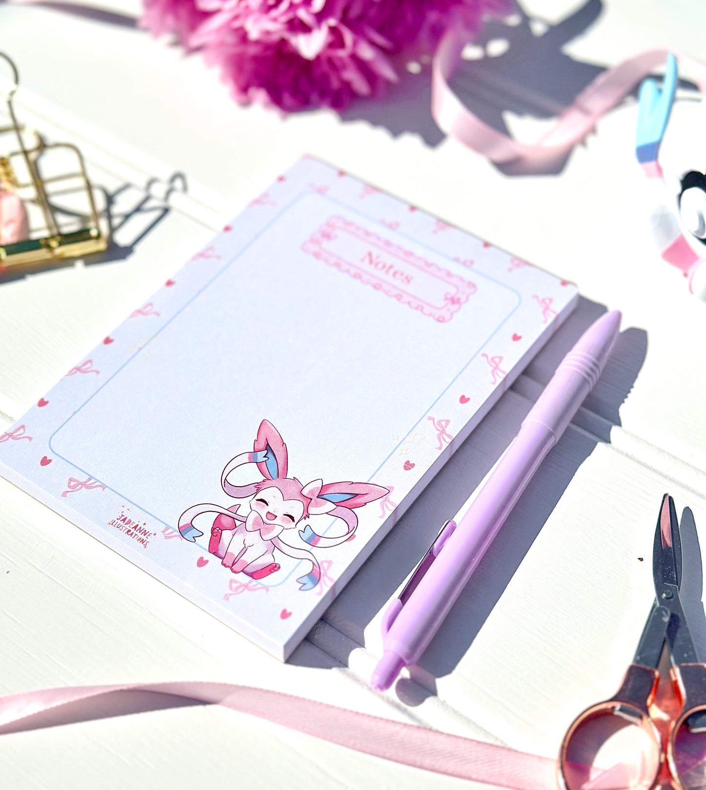 Notepad with a cute Sylveon character design, pen, and scissors on a light surface.