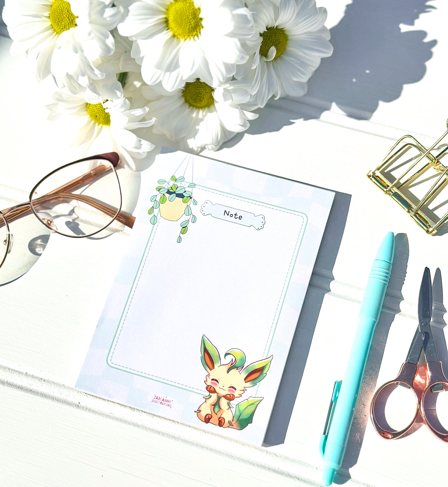 Notebook with cute leafeon pokemon illustrations, glasses, pen, scissors, and flowers on a white surface