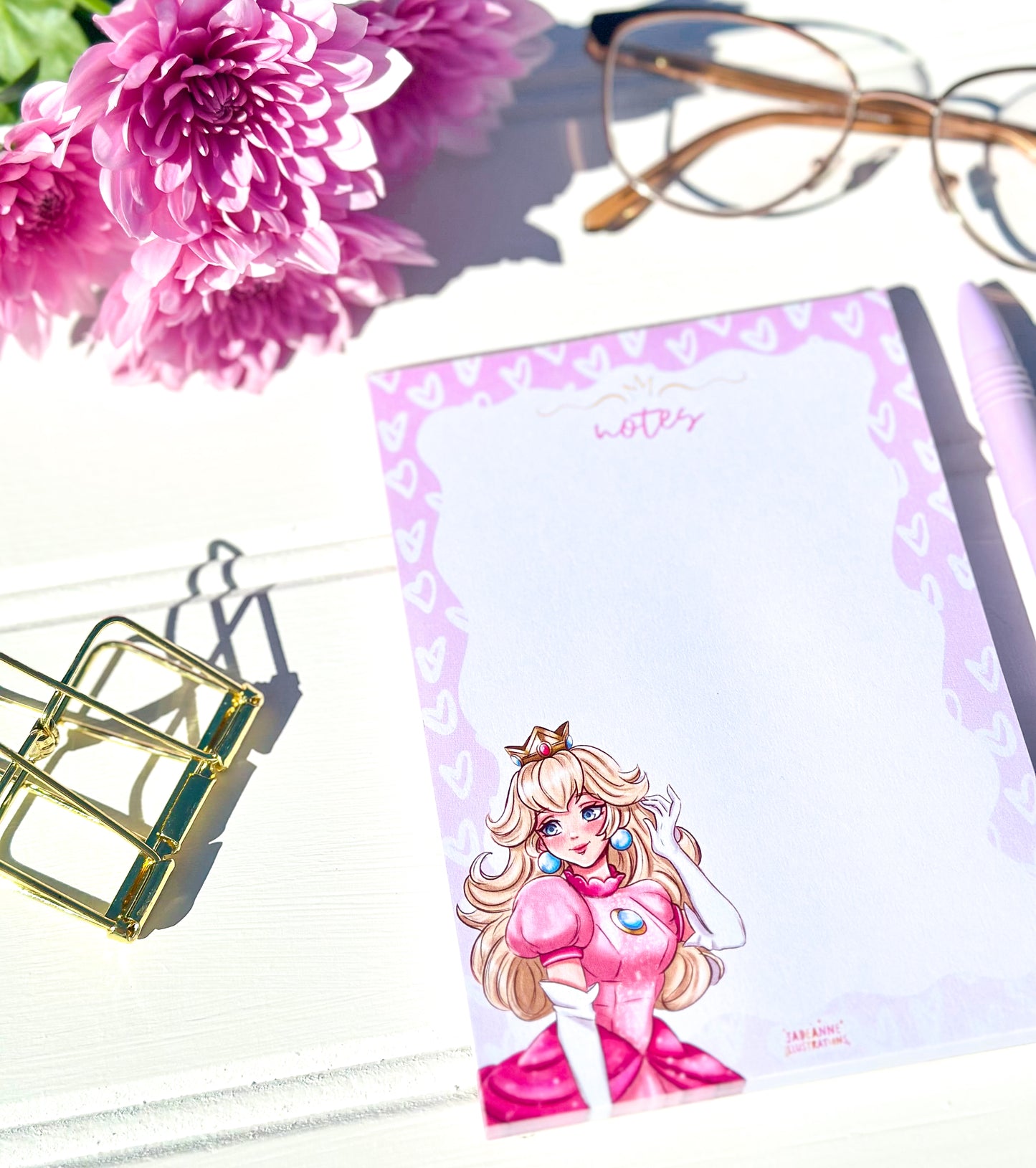 Notepad with a princess character design, pink flowers, glasses, and a pen on a white surface
