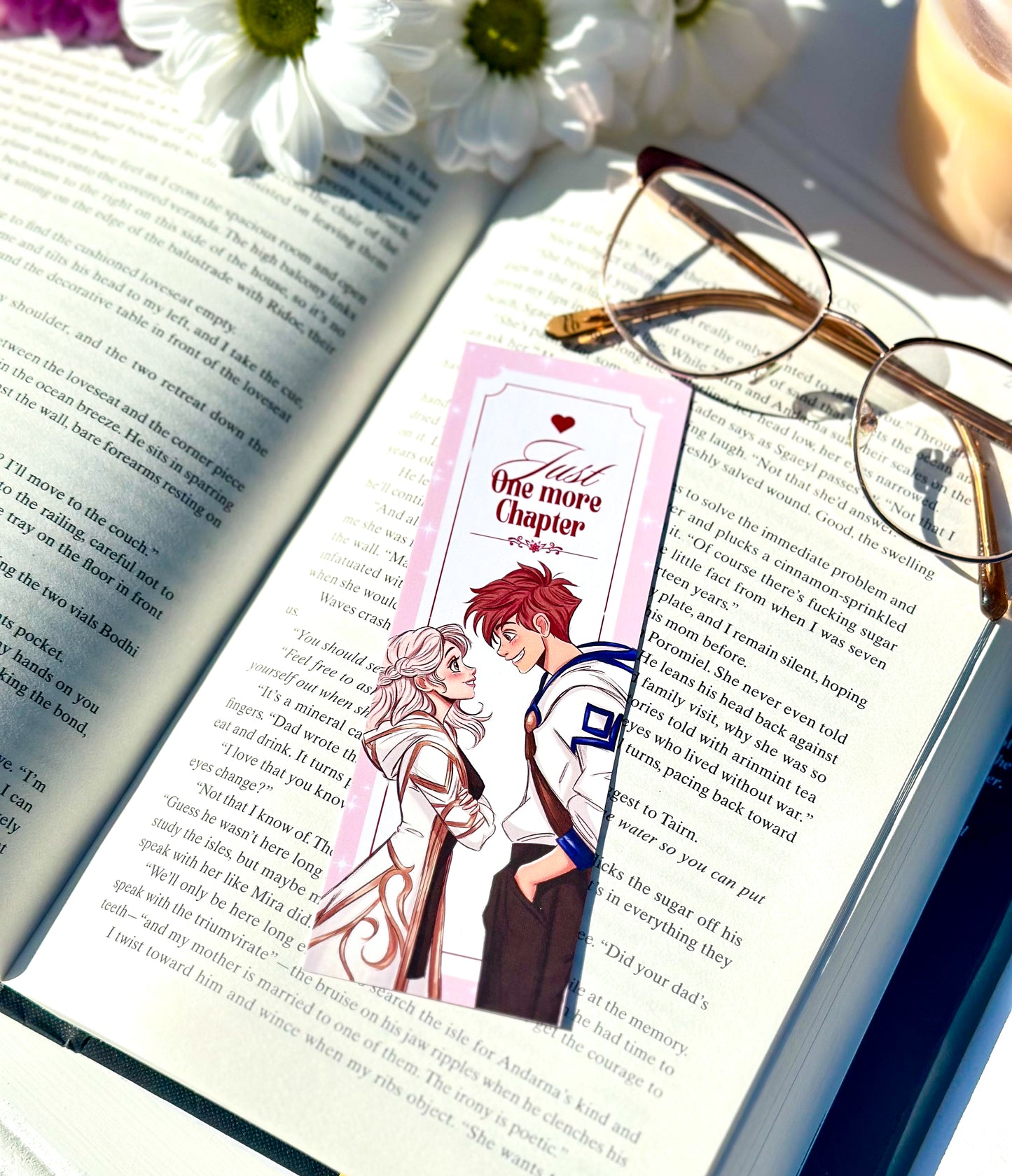 Open book with a bookmark featuring anime-style romantic couple characters, glasses on the page, and flowers in the background.
