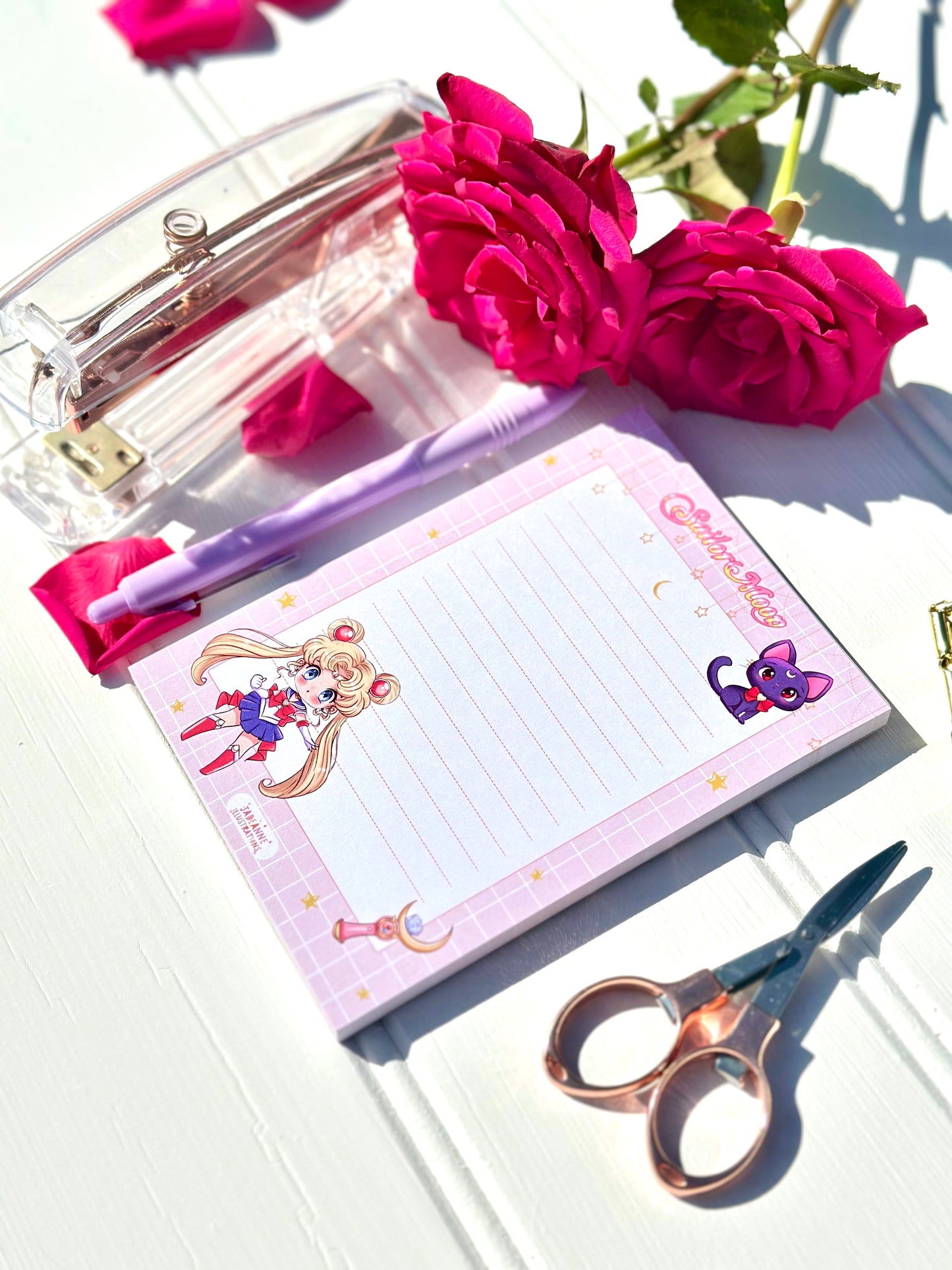 Notepad with Sailor Moon design, pen, scissors, and flowers on a white surface