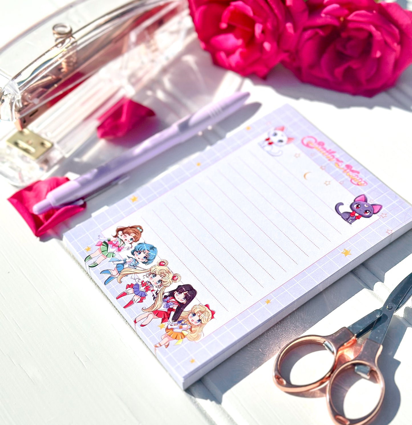 Notepad with chibi sailor moon characters, pen, and scissors on a white surface with pink flowers.