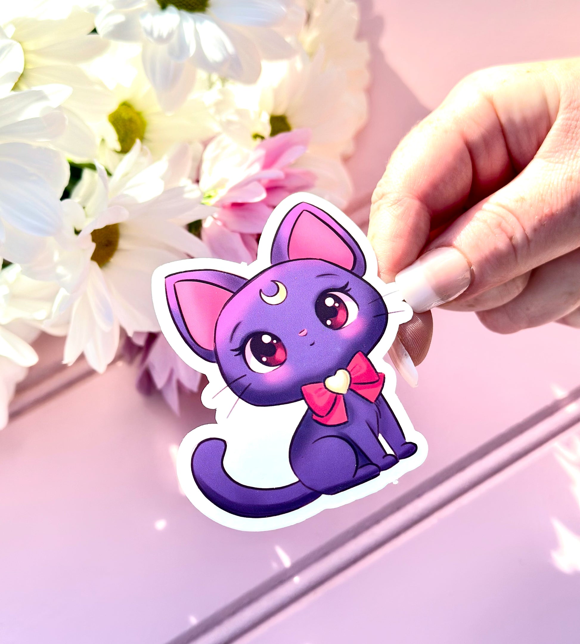 Hand holding Luna from sailor moon cartoon cat sticker with flowers in the background