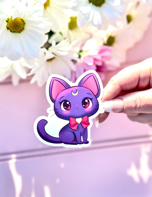 Hand holding a Luna cat-shaped  sailor moon sticker with pink accents against a floral background