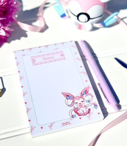 Notebook with a cute pokemon sylveon character design on a white surface with a pen and flowers.