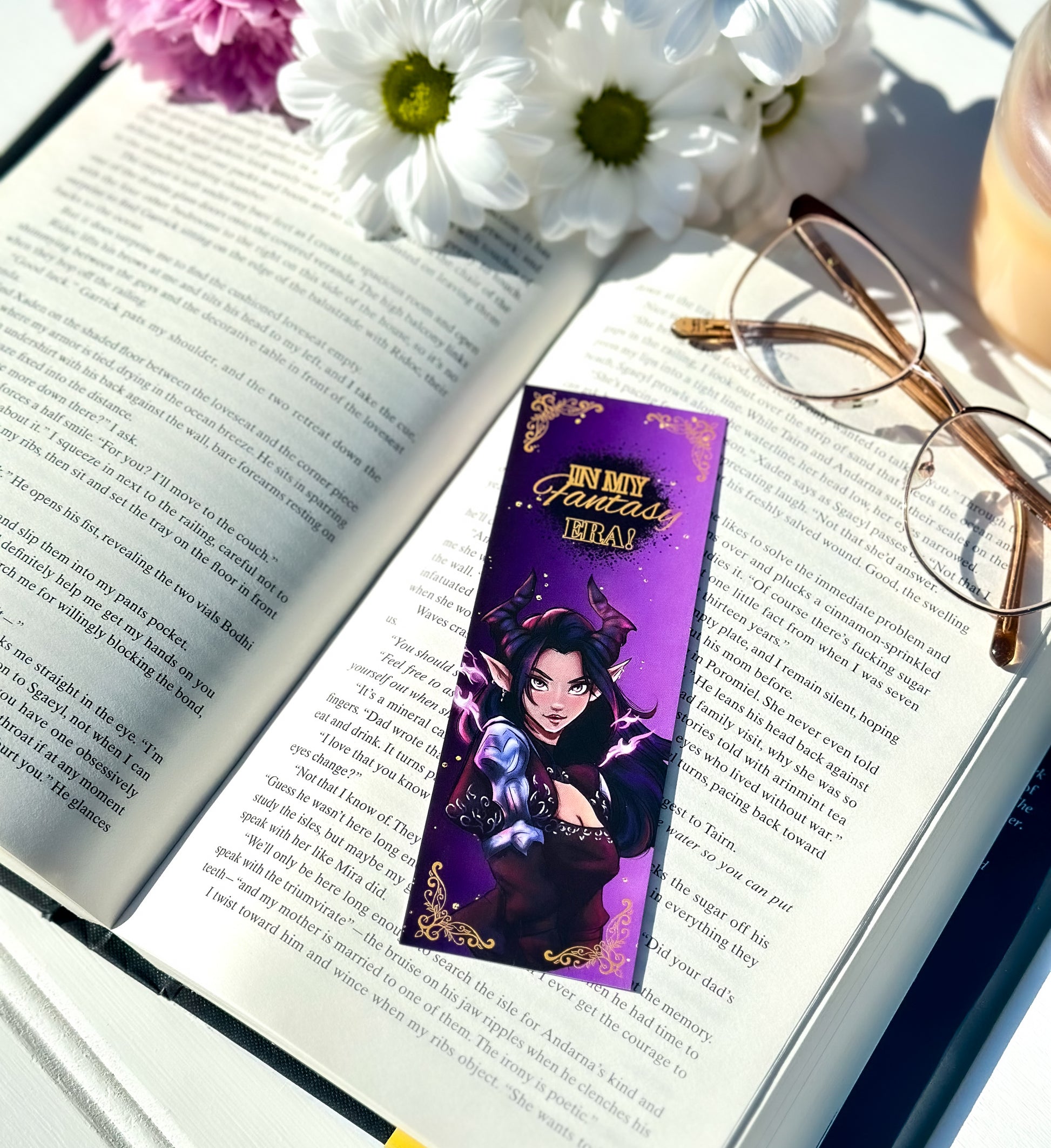 Open book with a purple bookmark featuring a dark fae character, surrounded by flowers and glasses.