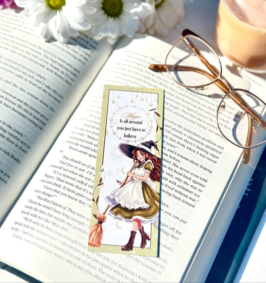 Open book with a bookmark featuring a witch, glasses, and flowers on a sunny surface.