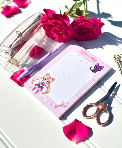 Notebook with Sailor Moon design, pen, and scissors on a white surface with pink flowers.