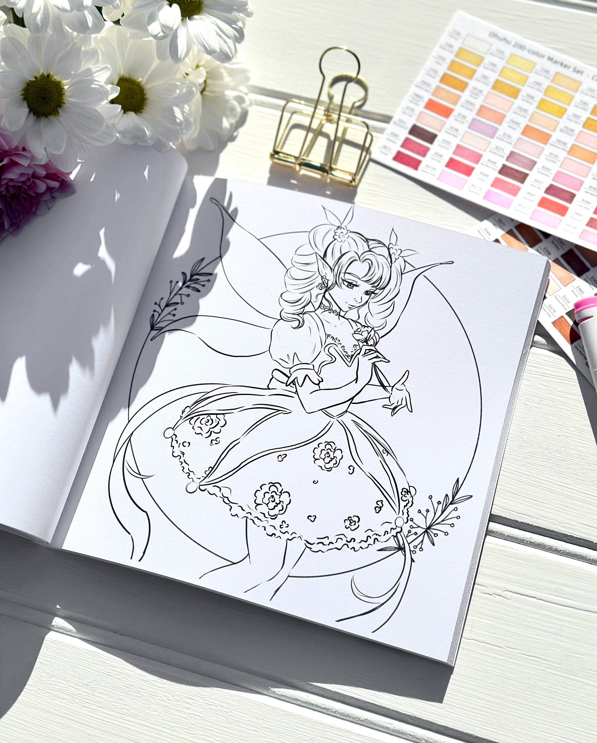 Colouring book page with a fairy design, flowers, and colour swatches on a white surface.