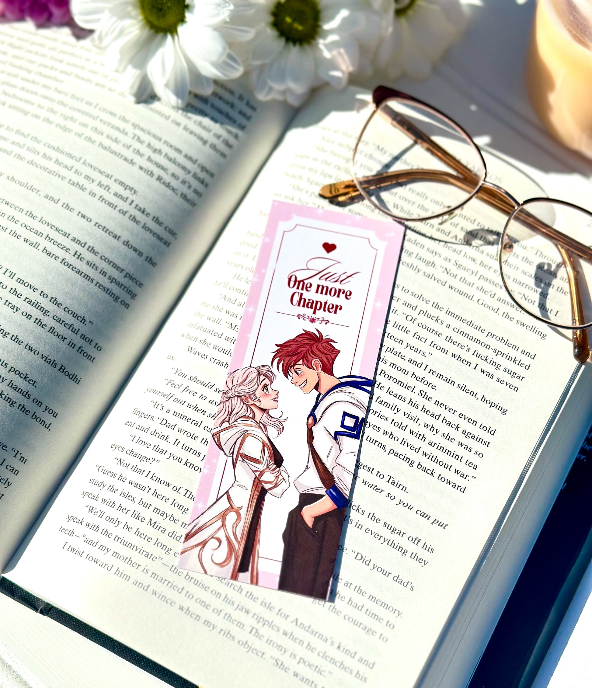 Open book with a bookmark featuring anime-style romantic couple characters, glasses on the page, and flowers in the background.